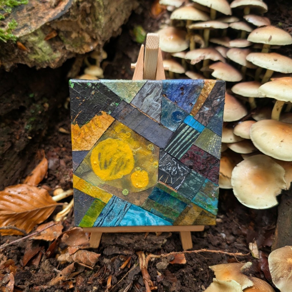 Photograph of a small, mixed media, abstract collage on canvas, featuring organic and geometric textures and patterns in a varied colour palette. The artwork is standing on a mini easel next to a cluster of pale fungi behind to the right. The easel is standing on the bare earth with a few brown, fallen leaves scattered around. A portion of a fallen log with green moss is visible top left.