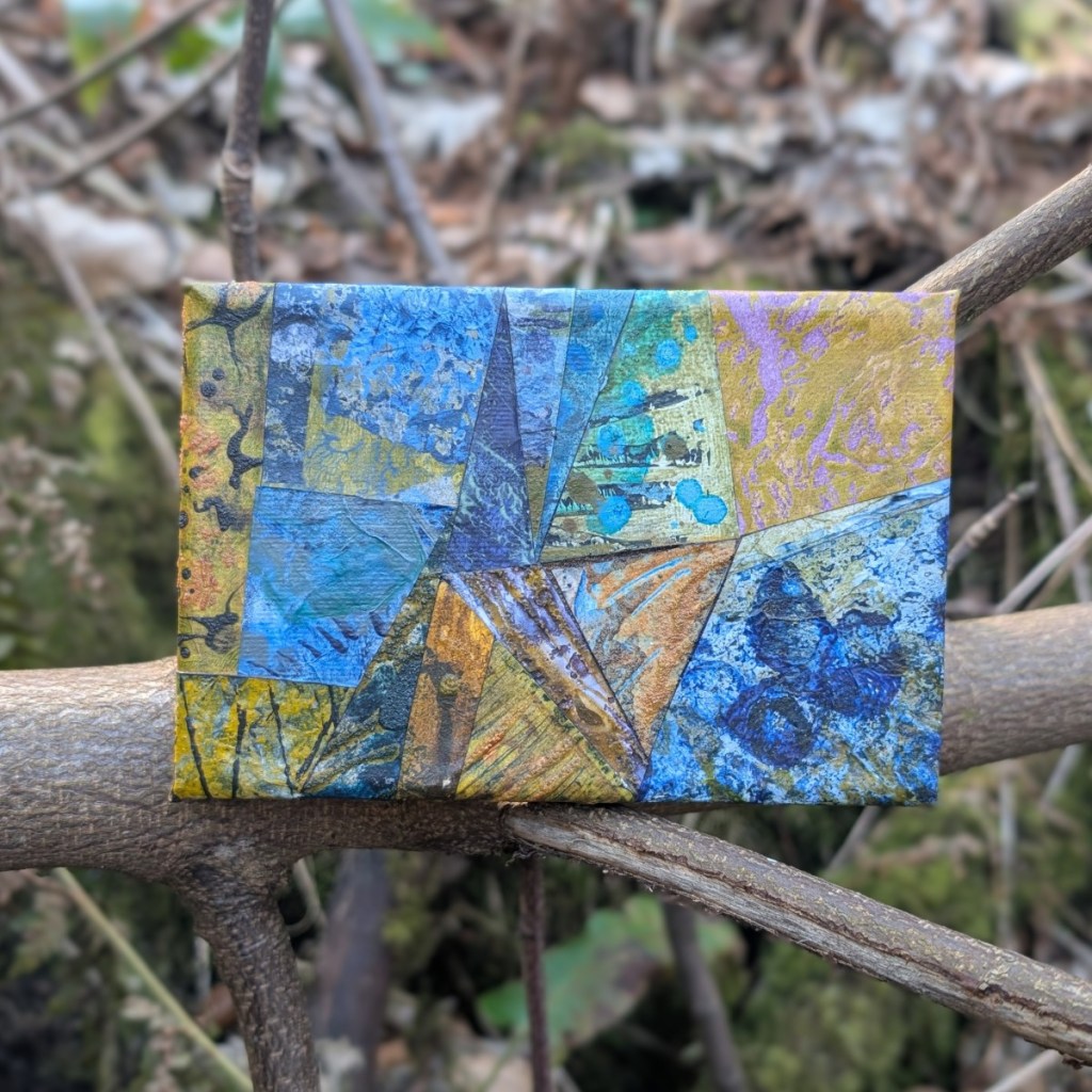 A photograph of a small, mixed media, abstract collage on canvas, featuring organic and geometric textures in a colour palette of blues, yellows and pale greens. The artwork is propped against bare branches with undergrowth of fallen leaves and green foliage visible out of focus behind.