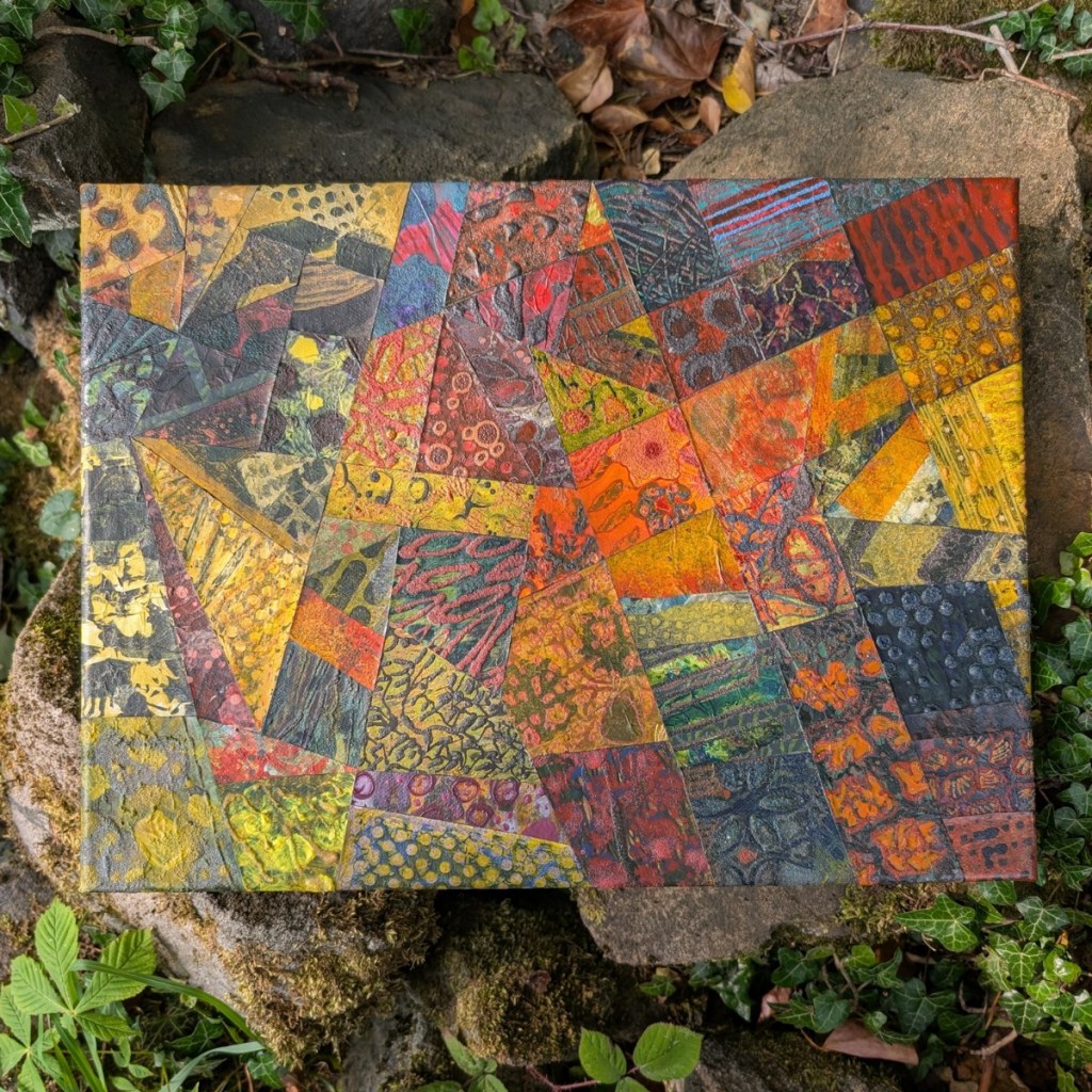 A photograph of a mixed media, abstract collage on canvas, featuring organic and geometric textures and patterns in a varied colour palette. The artwork is in landscape orientation, lying flat on top of mossy rocks and undergrowth including dried leaves, ivy and brambles.