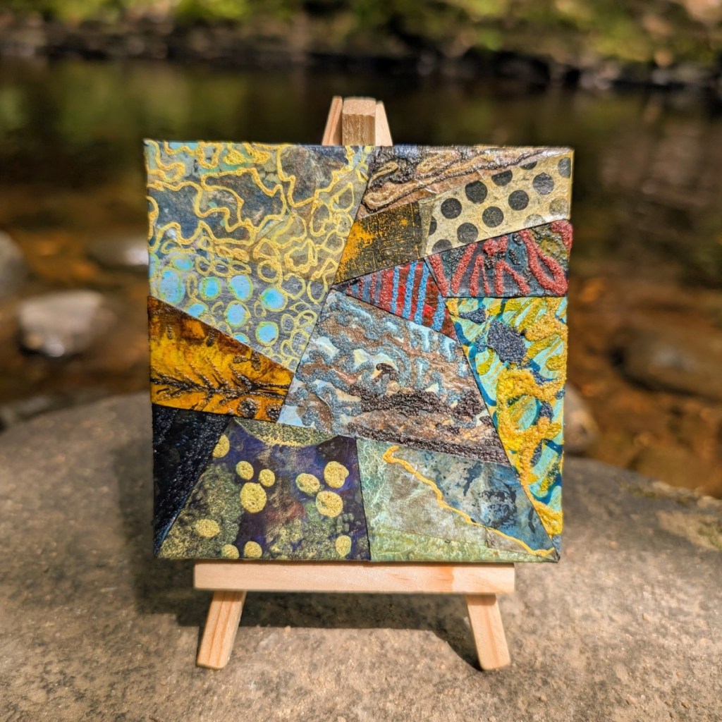 Photograph of a small, mixed media, abstract collage on canvas, featuring organic and geometric textures and patterns in a varied colour palette. The artwork is standing on a mini easel on a rock. The stream and opposite bank are visible behind, out of focus.