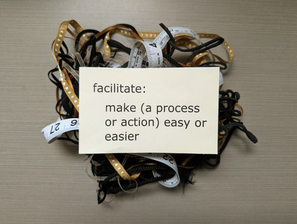 A photograph of a yellow sticky note displaying printed text which reads 'facilitate: make (a process or action) easy or easier'. The note is on top of a heap of ribbon, laces and cord all tangled up with a sewing tape measure. There is a mid grey tiled surface below.
