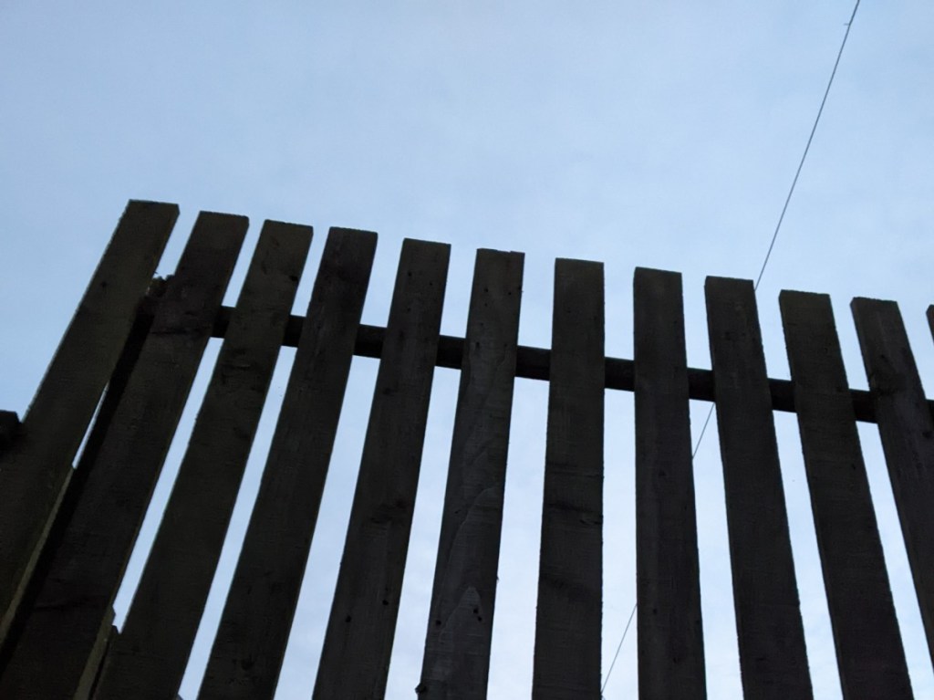 A photograph of a wooden fence silhouetted starkly against a pale blue sky. Eleven uprights and a single crossbar rise at a slight angle to the viewer, sloping left to right. A telephone line is also visible, slanting down from right to left on the right hand side of the image.