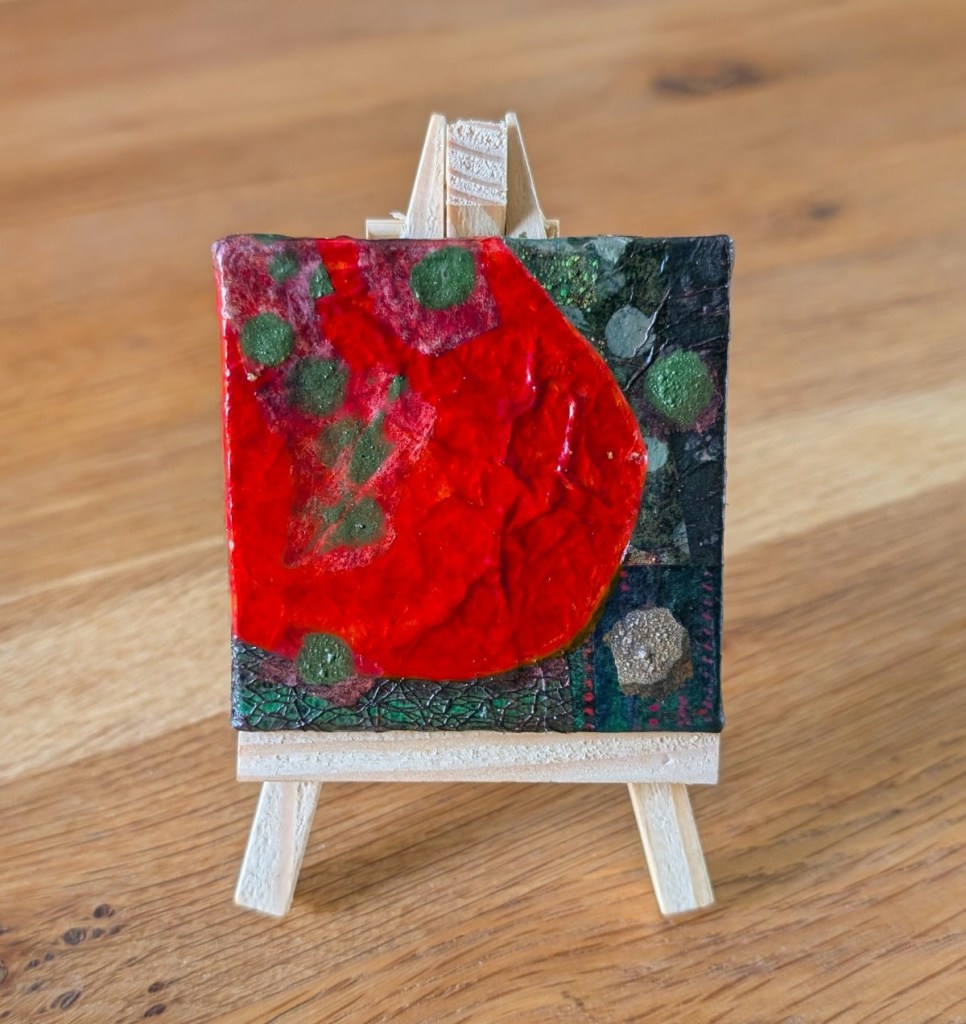 A small, abstract collage artwork displayed on a wooden mini easel with the grain of a wooden table behind. The collage features organic shapes and textures in a colour palette of reds and greens.
