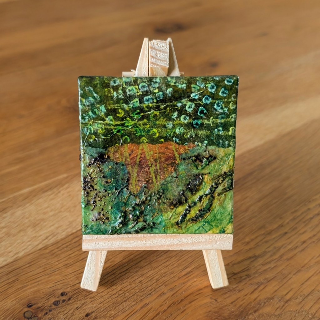 A small, abstract collage artwork displayed on a wooden mini easel with the grain of a wooden table behind. The collage features organic shapes and textures in a colour palette of browns and greens.