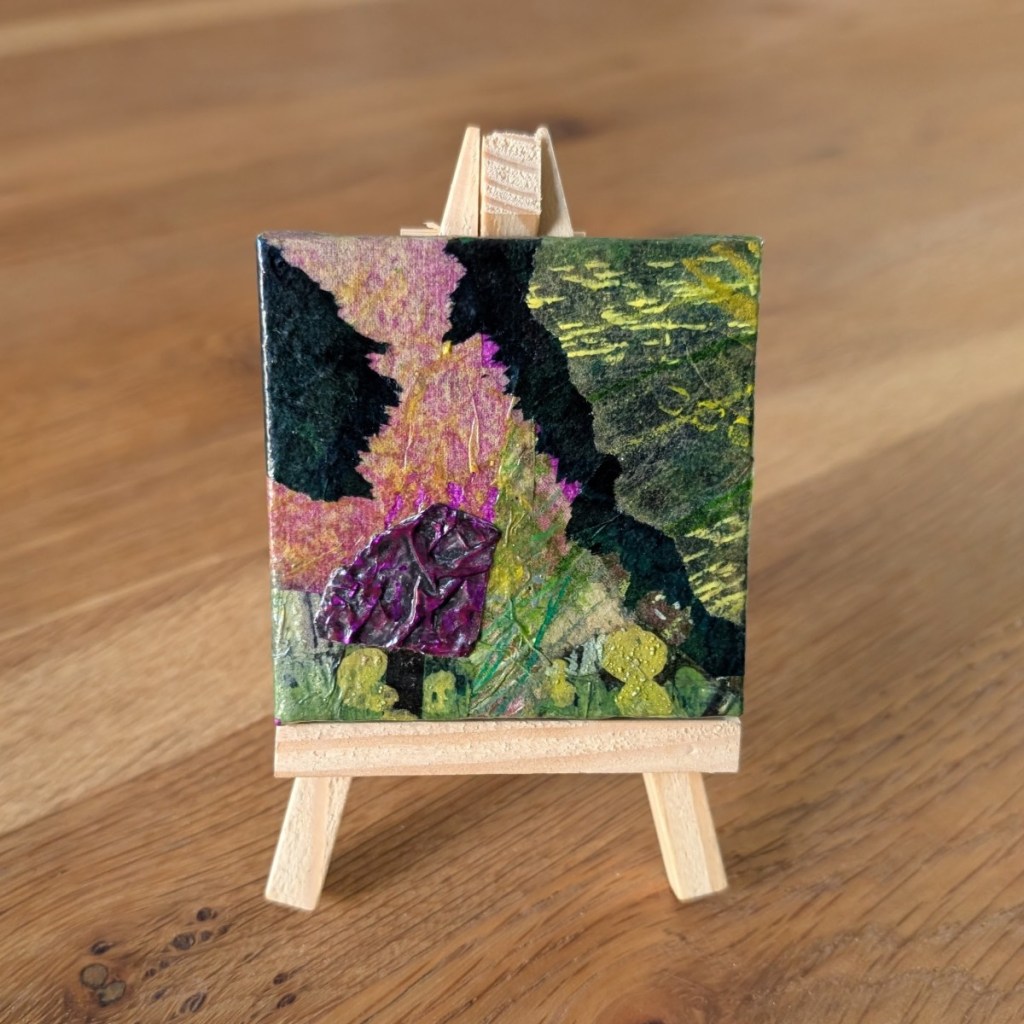A small, abstract collage artwork displayed on a wooden mini easel with the grain of a wooden table behind. The collage features organic shapes and textures in a colour palette of purples and greens.
