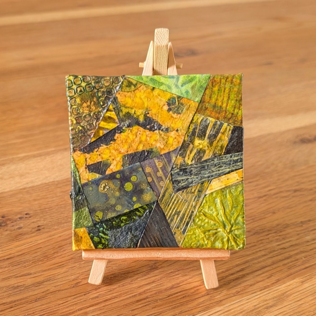 A small, abstract collage artwork displayed on a wooden mini easel with the grain of a wooden table behind. The collage features organic shapes and textures in a colour palette of greens and greys.