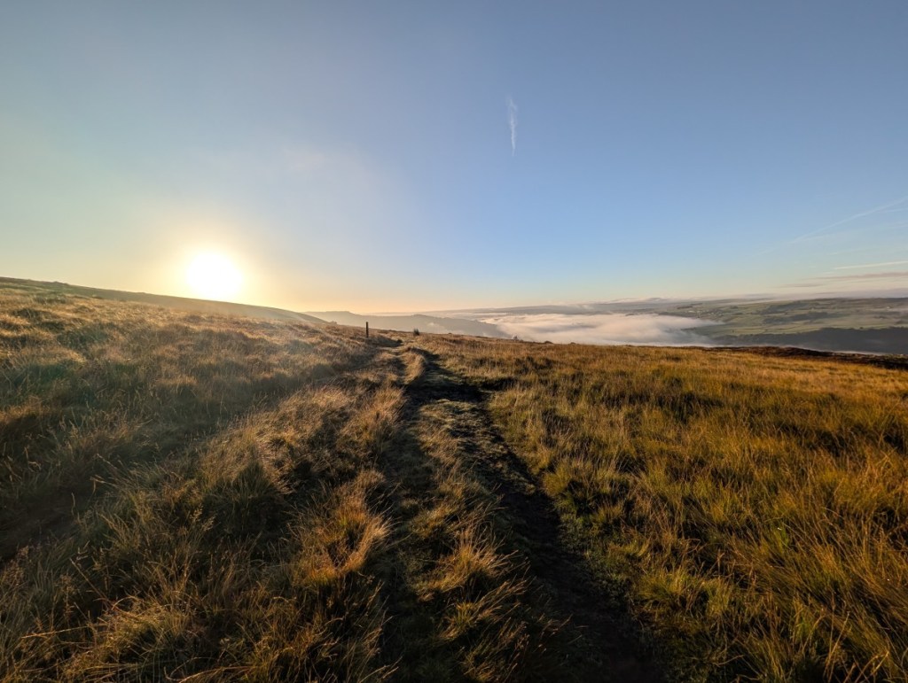 Photograph of the sun rising above Midgley Moor. The foreground features moorland grasses and a grassy track leading straight ahead. Cloud is gathered in the distant valley below. The sky is pale blue and slightly hazy.