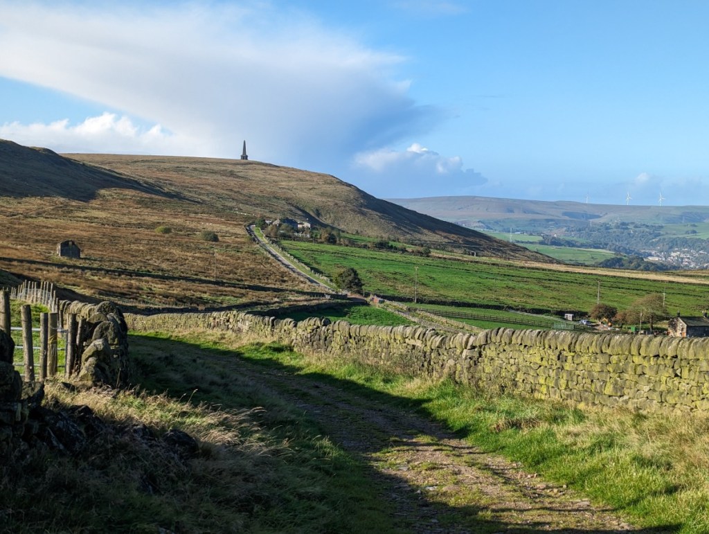 A morning photograph of the Calderdale landscape with Stoodley Pike Monument visible on a fell top in the middle distance. In the foreground a lane leads down to the left, flanked by dry stone walls. The landscape is a mix of moorland, farmland and woodland with wind turbines visible on the distant fell tops. The sky is blue with a large bank of grey and white cloud behind the monument.