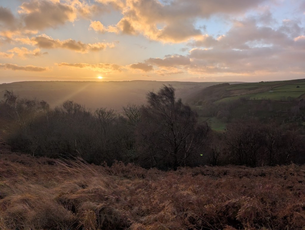 A photograph of the view towards the east from Broadhead Clough early on an autumnal morning. The sun is rising behind a distant fell casting a golden light across the landscape.