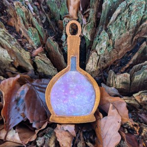 A mixed media charm in the shape of a bottle featuring a coppery gold outline with a semi-translucent central section. The bottle contains a bubbly-looking substance with a red tint. The charm is set against the textures of a tree stump full of autumnal leaves.