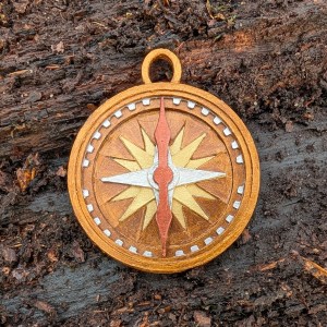 A mixed media charm for a bracelet in the shape of a compass. The charm has a coppery gold finish with details picked out in silver, pale gold and copper. The copper need points north and the compass is set against the bark of a fallen log.