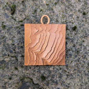 Photograph of a square mixed media charm for a bracelet, set against a grey stony background. The charm has a coppery gold finish and features a series of layers building up a representation of the contours on an OS map.