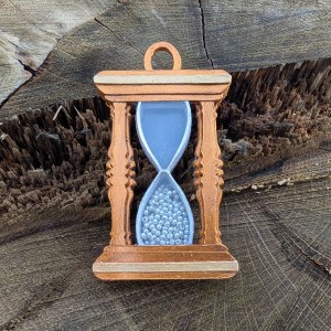 A mixed media charm for a bracelet in the shape of an hourglass. The silhouette of the frame has a coppery gold finish with pale gold horizontal accents top and bottom. Inside is set a translucent section with silvery edges, representing the glass. The bottom half is filled with tiny silvery beads representing fallen sand. The charm is set against the textures of a tree stump.