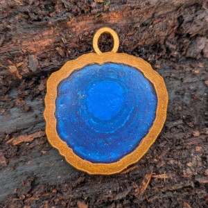 Photograph of a mixed media charm for a bracelet representing a pool of water. The charm is roughly round in shape and its borders have a coppery gold finish. Inside is a semi-translucent mid blue section with layered details suggesting depth. The charm is set against the textures of a fallen log.