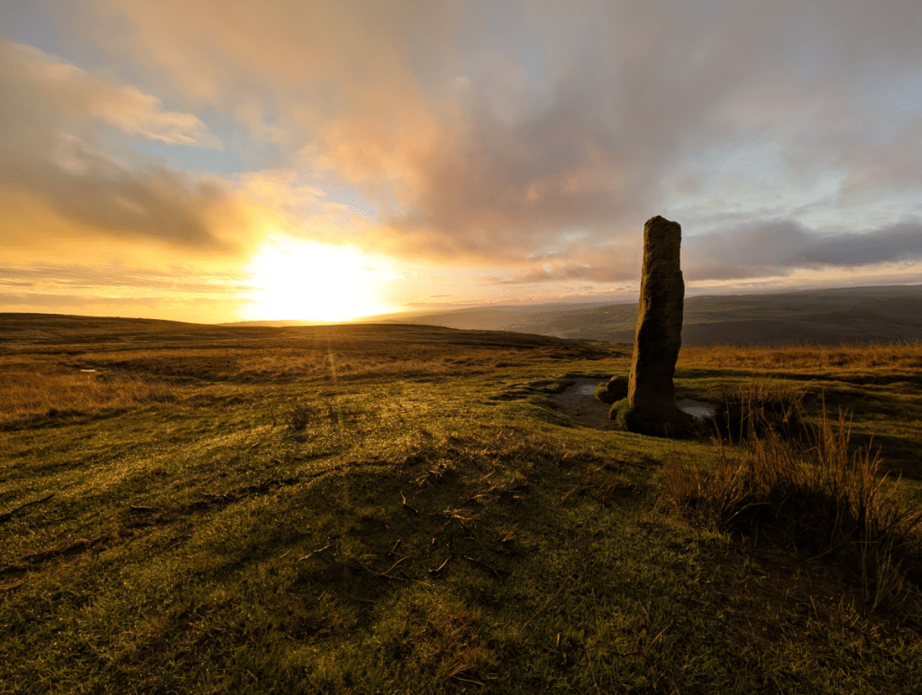 Photograph of an autumnal sunrise featuring the Churn Milk Joan standing stone on Midgley moor. The stone, to the right of the image, is partially lit by the sun, to the left of the image, as it begins to rise above the horizon in a dramatic blaze of white gold.