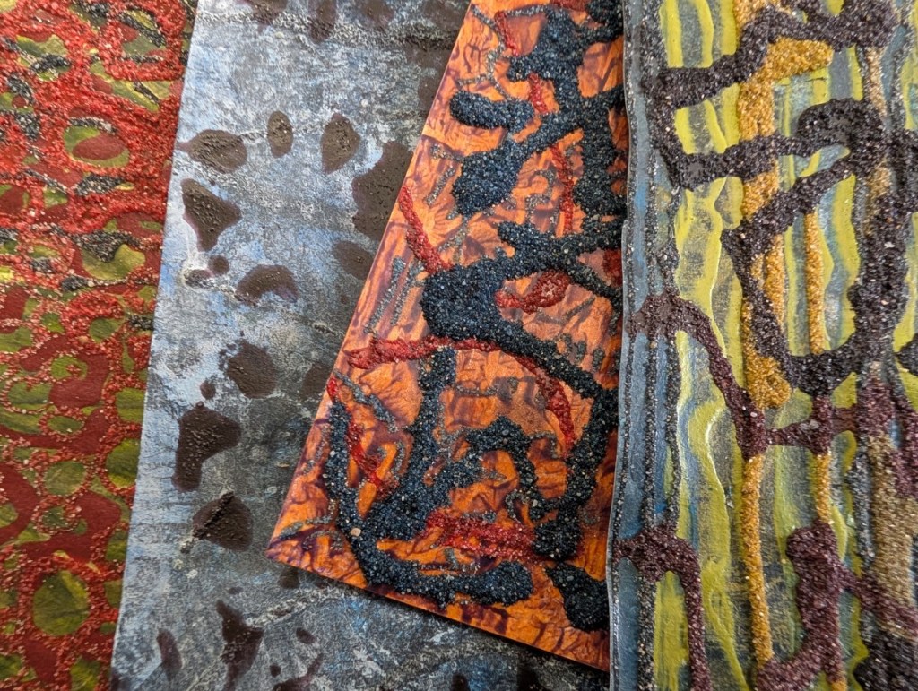 A photograph of assorted mixed media work on paper featuring  mark-making in dark, gritty textured paint against contrasting colours.