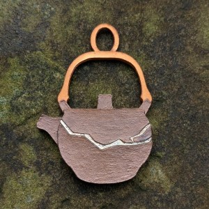 A mixed media charm for a bracelet in the shape of a Japanese-style teapot, in profile with the spout to the left. The charm has a coppery gold finish on the teapot handle and the main body is brown with a metallic lustre. There is a  horizontal crack across the body of the pot, with a gold fill. The charm is set against stone with mosslike textures in shades of green.
