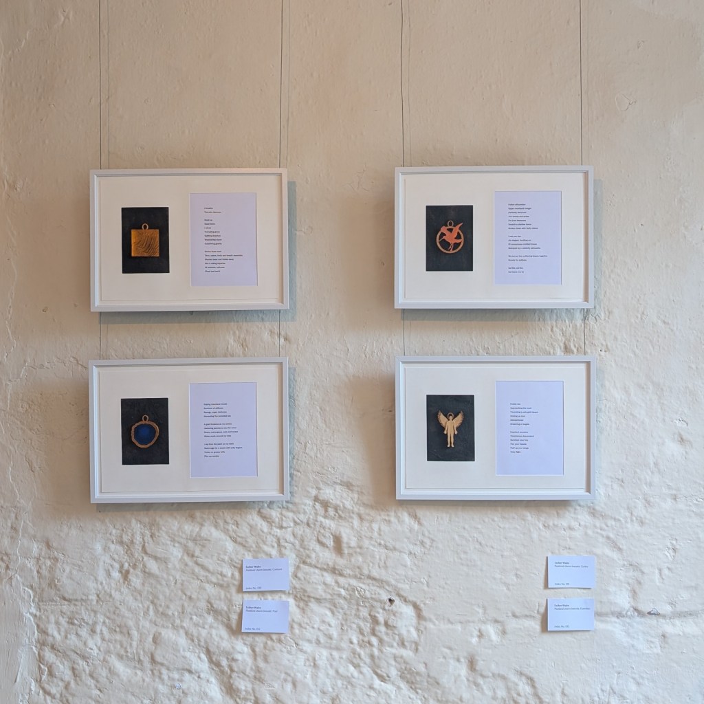 A photograph of four Peatland Charm Bracelet pieces installed for display at Gibson Mill. Each one comprises a mixed media charm set against a dark rectangular background, mounted side by side with its companion piece of writing with the charm to the left. These are displayed in four white frames, hung two by two in landscape orientation against an off-white stone wall.