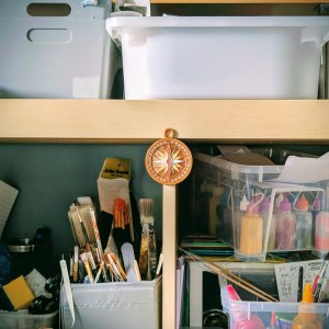 A mixed media charm for a bracelet in the shape of a compass. The charm has a coppery gold finish with details picked out in silver, pale gold and copper. The copper need points north. The compass is mounted on the front of a storage unit containing creative materials including paints, brushes and storage boxes.
