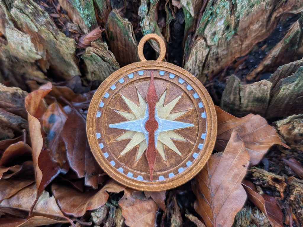 A mixed media charm for a bracelet in the shape of a compass. The charm has a coppery gold finish with details picked out in silver, pale gold and copper. The copper need points north and the compass is set against the bark of a fallen log strewn with fallen leaves.