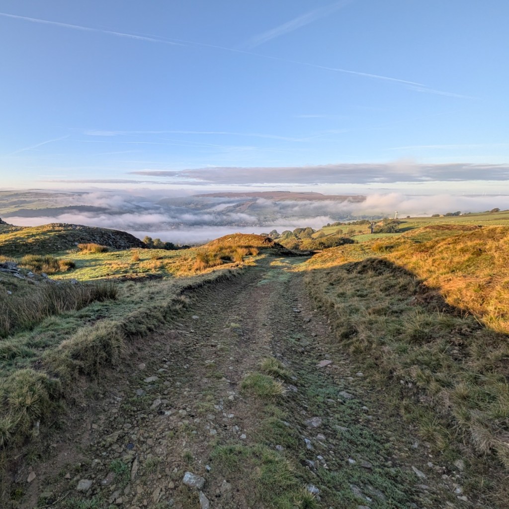 Photograph of a broad track leading forwards across Midgley moor. In the distance, clouds are scattered across the valley below and in front of the fells rising in the far distance. The silhouette of Stoodley Pike Monument is just visible on the horizon, above which there is blue sky. The morning sunlight is casting shadows across the foreground; some nearby areas of moorland, farmland and woodland are picked out in vibrant colour by the early light.