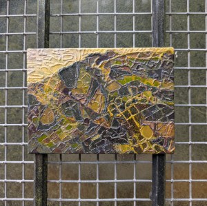 Photograph of a small, landscape-orientation mixed media artwork featuring organic mark-making in a colour palette of greens, greys, yellows and browns.

The artwork is set against a two vertical, black metal struts in front of grey wire mesh protecting an opaque glass window.