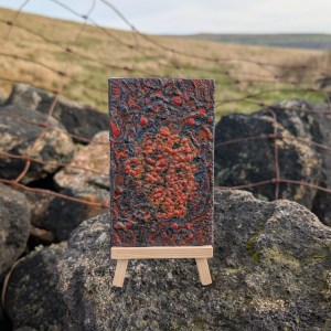 Photograph of a small, portrait-orientation mixed media artwork featuring organic mark-making in a colour palette of oranges and greys. The artwork is supported by a mini easel on a dry stone wall. Behind, out of focus, is a half-fallen section of rusted metal fencing beyond which rough grassland extends towards distant fell tops under a grey-blue sky.