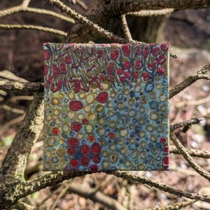 Photograph of a small, square mixed media artwork featuring organic mark-making in a colour palette of greens, reds and blues. The artwork is set against bare conifer branches.