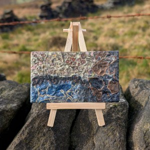 Photograph of a small, landscape-orientation mixed media artwork featuring organic mark-making in a colour palette of blues, greys, pinks and browns. The artwork is supported by a mini easel on top of a dry stone wall. Two rusted strings of barbed wire run horizontally behind. Grasses and sections of wall are visible in the background, out of focus.