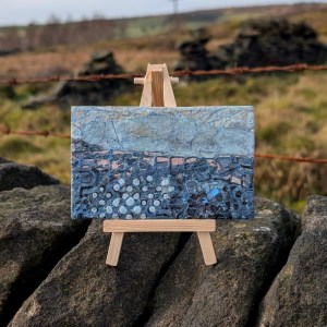 Photograph of a small, landscape-orientation mixed media artwork featuring organic mark-making in a colour palette of blues and greys. The artwork is supported by a mini easel on top of a dry stone wall. Two rusted strings of barbed wire run horizontally behind. Grasses and sections of wall are visible in the background, out of focus.
