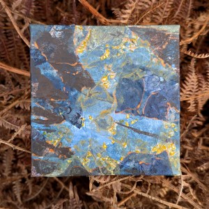 Photograph of a small, square mixed media artwork featuring organic mark-making in a colour palette of yellows, greys and blues. The artwork is set against bronze bracken fronds.