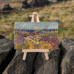 Photograph of a small, landscape-orientation mixed media artwork featuring organic mark-making in a colour palette of moorland shades. The artwork is supported by a mini easel on top of a dry stone wall. Two rusted strings of barbed wire run horizontally behind. Grasses and sections of wall are visible in the background, out of focus.