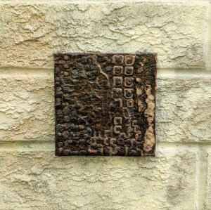 Photograph of a small, square mixed media artwork featuring organic mark-making in a colour palette of browns and yellow-greys. The artwork is set against a brick wall covered in off-white spray paint with a hint of yellow.