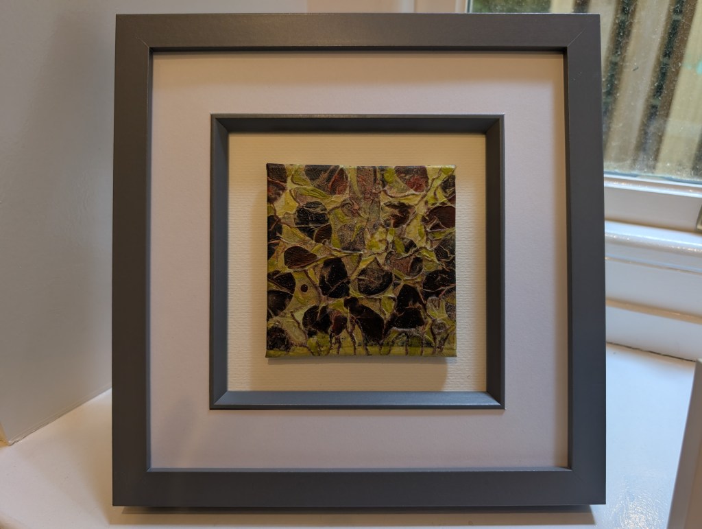 Photograph of a small, square mixed media artwork featuring organic mark-making in a colour palette of yellows and deep reds. The artwork is mounted in a square frame, set against a warm white background. The inner mount and outer edges of the frame are mid grey.