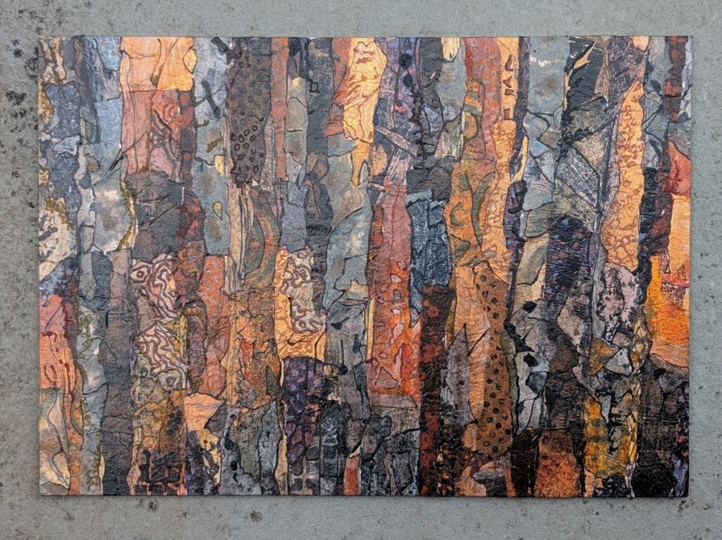 Photograph of a landscape-orientation mixed media artwork featuring organic mark-making evocative of textures of tree bark in a colour palette of golds, oranges, dark browns and greys. The artwork is set against a pale grey stone background.