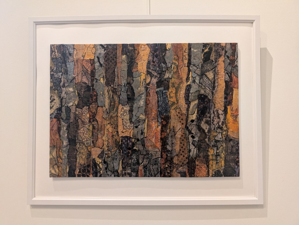 Photograph of a landscape-orientation mixed media artwork featuring organic mark-making evocative of textures of tree bark in a colour palette of golds, oranges, dark browns and greys. The artwork is mounted against a warm white background inside a white frame, hung on a white wall.