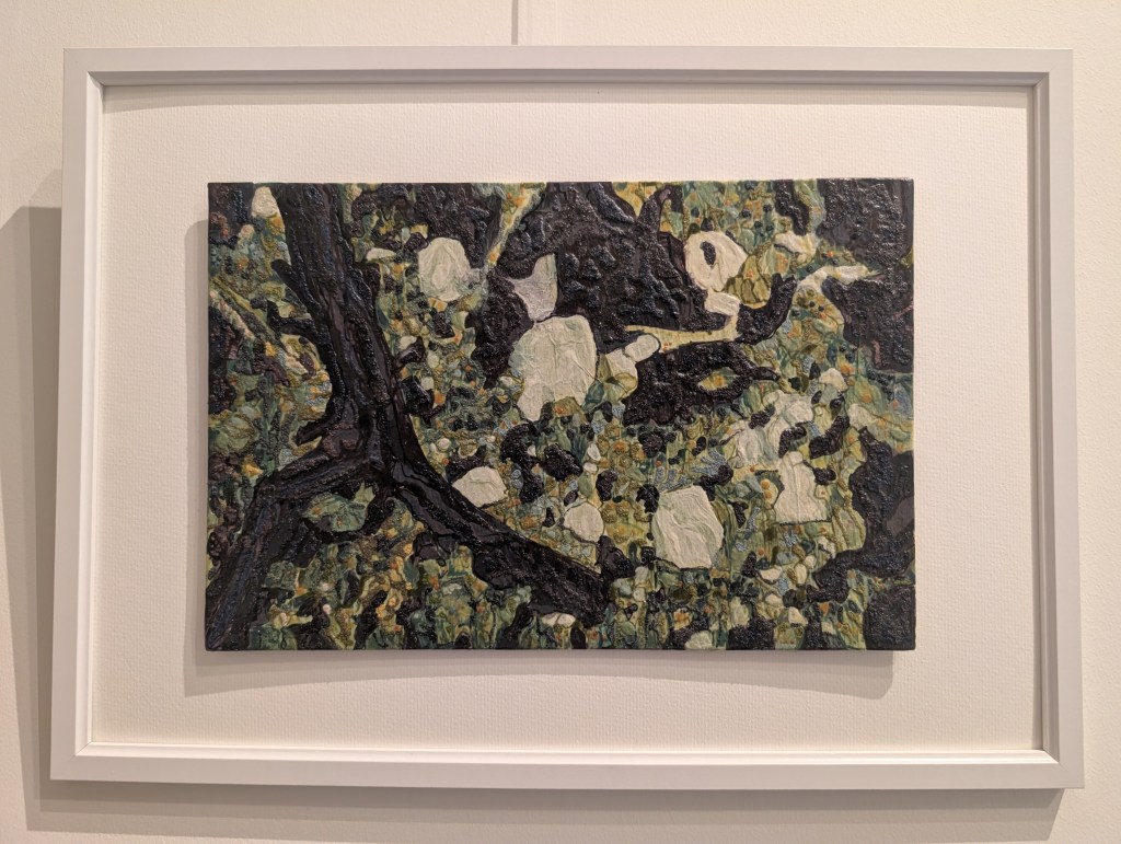 Photograph of a small, landscape-orientation mixed media artwork featuring organic mark-making evocative of branches and greenery in a colour palette of greens, dark browns, yellows and blues. The artwork is mounted against a warm white background inside a white frame, hung on a white wall.
