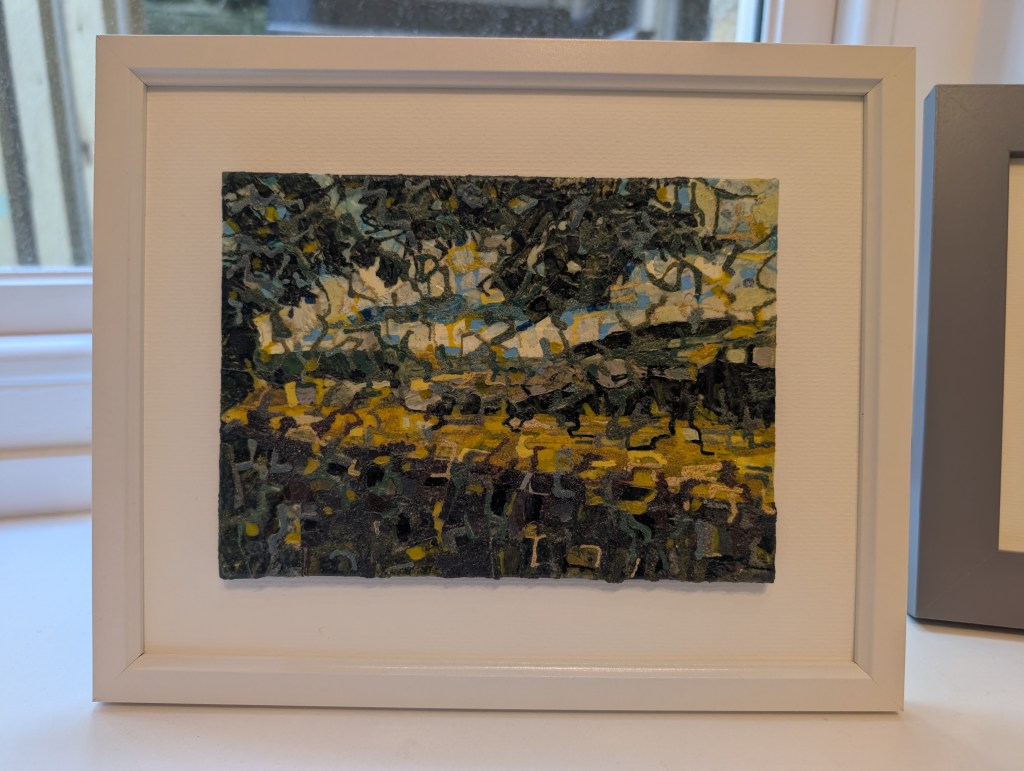 Photograph of a small, landscape-orientation mixed media artwork featuring organic mark-making in a colour palette of greens, near-blacks, yellows and blues. The artwork is mounted against a warm white background inside a white frame, standing on a windowsill.