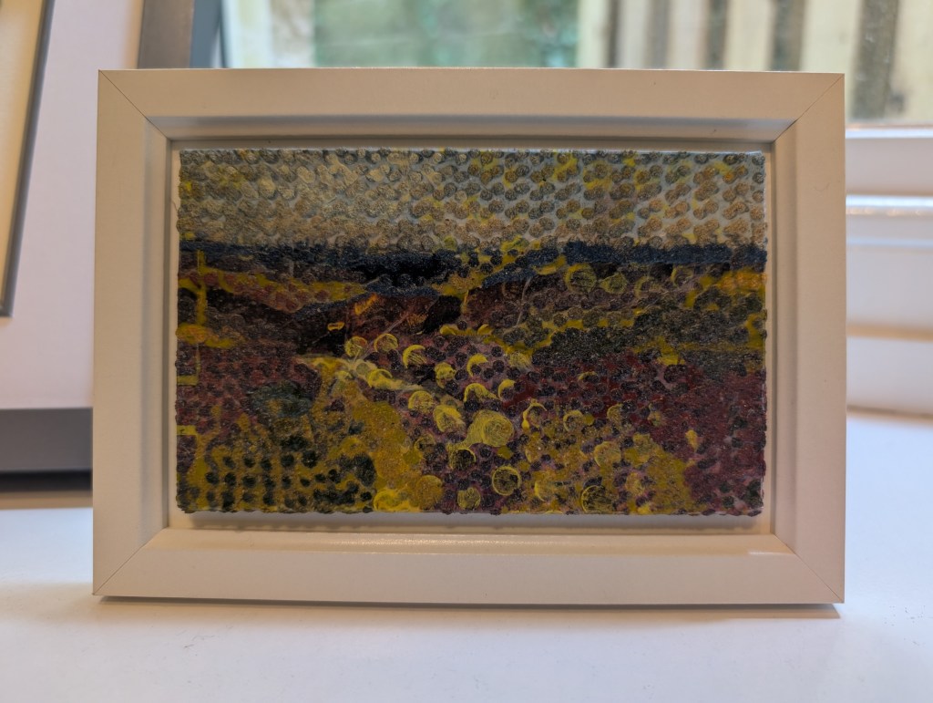 Photograph of a small, landscape-orientation mixed media artwork featuring organic mark-making in a colour palette of moorland shades. The artwork is mounted against a warm white background inside a white frame, standing on a windowsill.